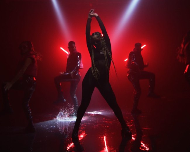 A dancer strikes a pose in the spotlight while three backup performers move in the background amidst dramatic lighting and reflections on a wet surface.