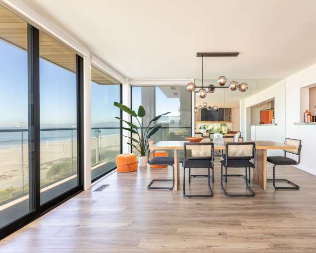 A dining area features a wooden table surrounded by black chairs, large windows showcasing a beach view, and modern light fixtures hanging overhead.