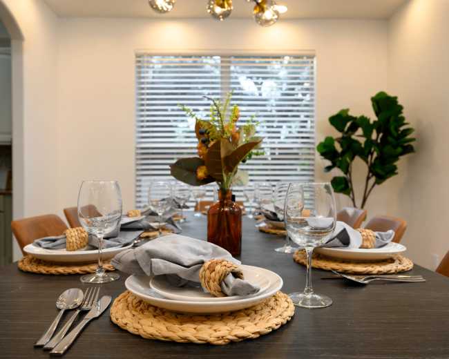 A dining table is set with glassware and neatly arranged plates, surrounded by chairs, and features a vase with flowers in a well-lit room.