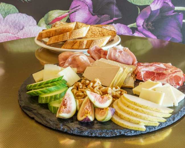 A platter of assorted cheeses, cured meats, fresh figs, and cucumbers is arranged on a slate board beside a plate of toasted bread slices.