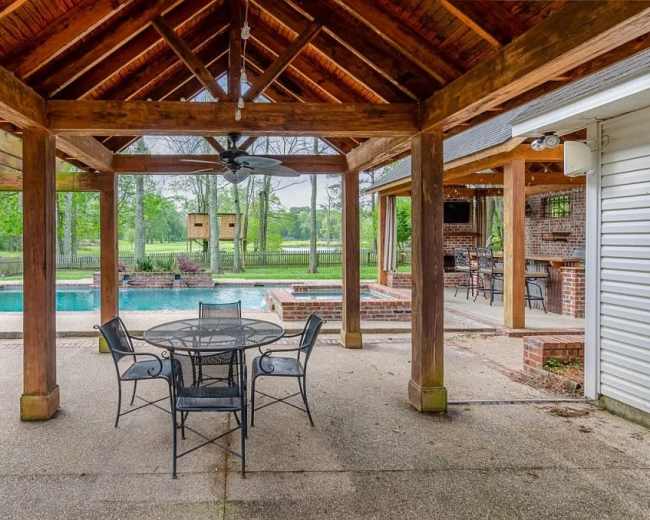 The image shows a covered outdoor patio area with a round dining table and chairs, overlooking a swimming pool and green landscape beyond.