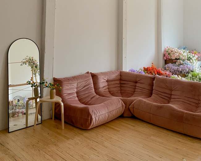 The room features a curved pink sofa, a small table with a plant, and a large mirror, with floral arrangements visible in the background and colorful garlands hanging from the ceiling.