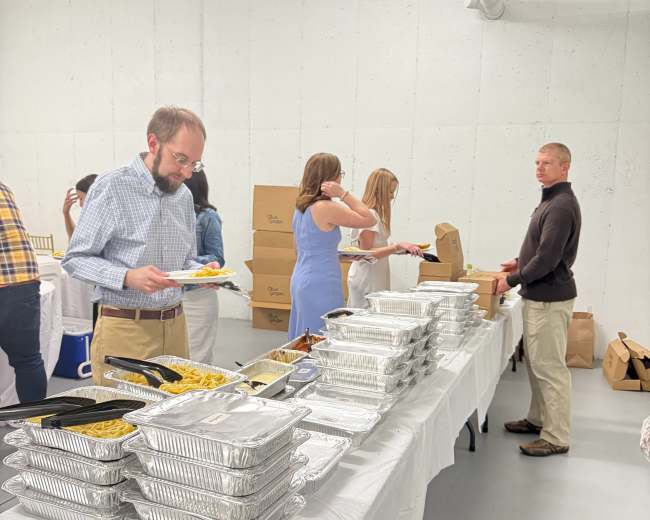 A group of people are serving themselves food from a buffet setup with multiple trays of dishes and takeaway boxes in a bright, spacious room.