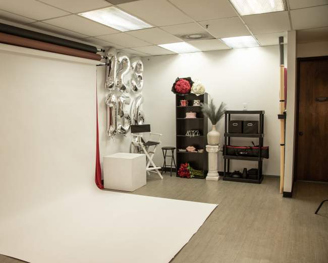 The image shows a photography studio equipped with a plain white backdrop, a styling area with decorative elements, and a shelving unit filled with props.