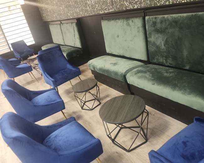 The image shows a modern interior of a café or lounge with green velvet benches, blue armchairs, and small round tables against a floral wallpaper background.