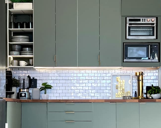 The image shows a modern kitchen featuring green cabinets, open shelves with kitchen items, and a round glass dining table with a gold base.