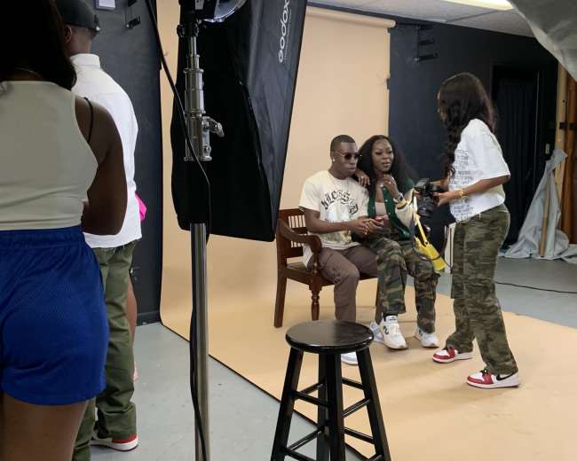 A photoshoot takes place in a studio with subjects posing on a chair against a backdrop, while a photographer and assistants manage lighting and setup.