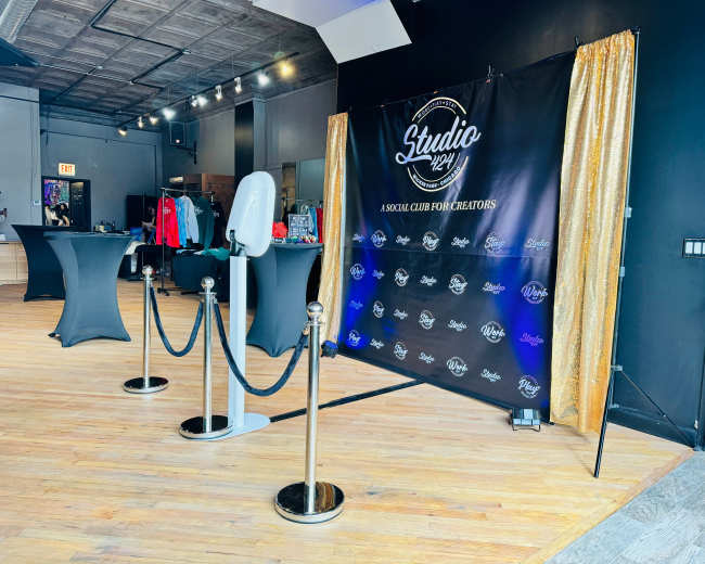 The image shows a spacious event space with a branded backdrop, black tables, and a clothing display against a dark wall.