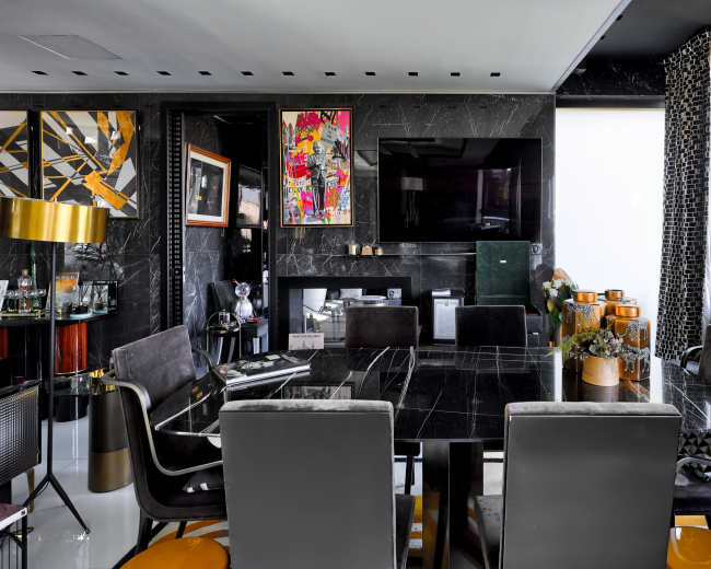 The image shows a modern dining area featuring a black marble table surrounded by dark chairs, with a large television mounted on the wall and colorful artwork displayed.