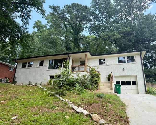 The image shows a light-colored, two-story suburban home with a garage, surrounded by trees and a small yard.