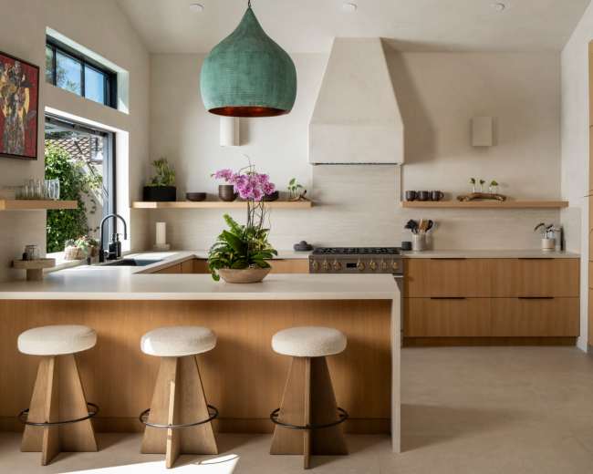 A modern kitchen features wooden cabinetry, a large island with three stools, and a pendant light above, accented by a countertop filled with plants and kitchenware.