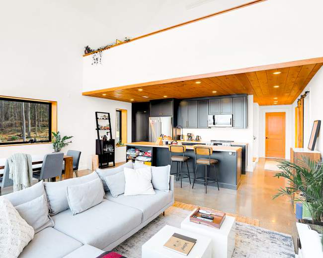 A modern living space featuring a light grey sectional sofa, a dining area with a table and chairs, and a kitchen with dark grey cabinetry and stainless steel appliances, all illuminated by natural light from large windows.