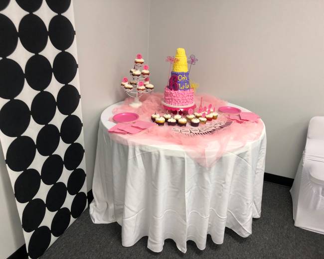A decorated table features a large cake topped with colorful icing and a tier of cupcakes, surrounded by pink plates and napkins.
