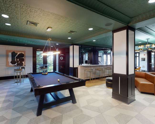 The image shows a modern lounge area featuring a billiards table, bar seating, and comfortable chairs under a decorative ceiling.
