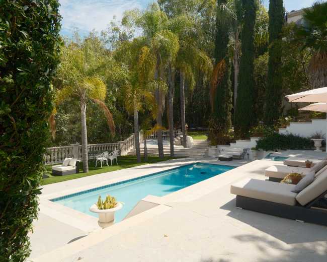 The image depicts a serene outdoor pool area surrounded by palm trees, lounge chairs, and stone pathways, with a view of a landscaped garden in the background.