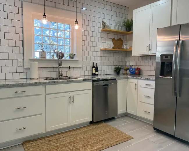 A modern kitchen features white cabinets, a stainless steel refrigerator, a dishwasher, and a window above the sink surrounded by subway tile.