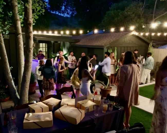 A group of people gathers and mingles under string lights in a backyard setting during the evening.