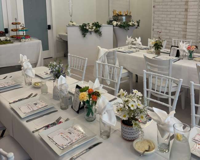 The image shows a neatly arranged dining area with white tables set with floral centerpieces, folded napkins, and place settings, complemented by decorative lighting and a buffet table in the background.