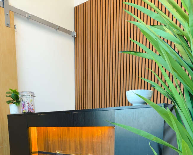 The image shows a modern reception desk with a wooden panel and illuminated sections, labeled "3A STUDIO," against a backdrop of wooden slats and indoor plants.