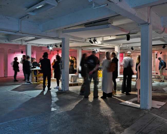 A crowd of people mingles in a spacious, dimly lit gallery featuring artworks and illuminated wall installations.