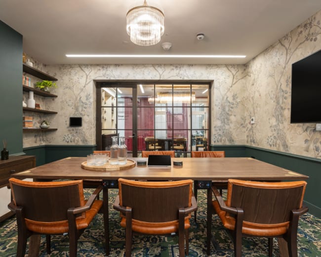 The image shows a conference room featuring a long wooden table surrounded by brown chairs, with floral wallpaper and a chandelier above.