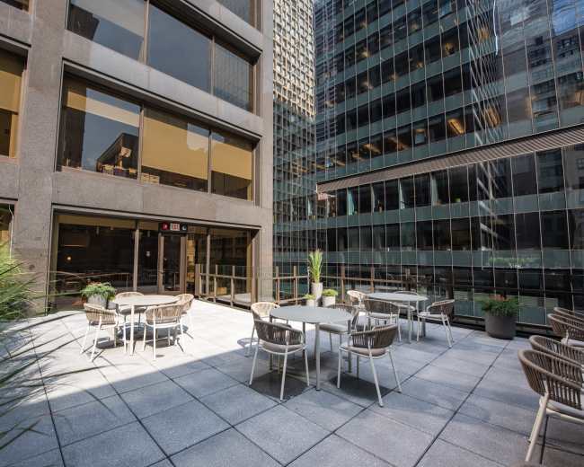 A concrete outdoor terrace features tables and chairs surrounded by tall office buildings.