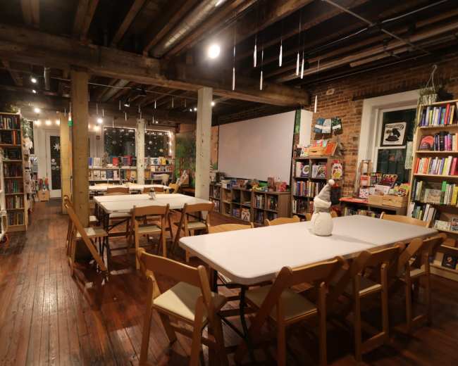 The image shows a cozy bookstore interior with wooden floors, bookshelves filled with various books, and several tables set up for reading or events.