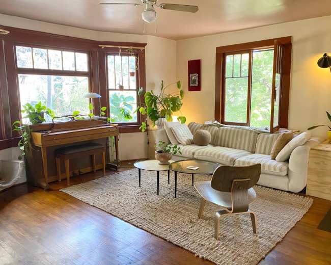 A cozy living room features a white sofa, a wooden piano, plants, and large windows allowing natural light to fill the space.
