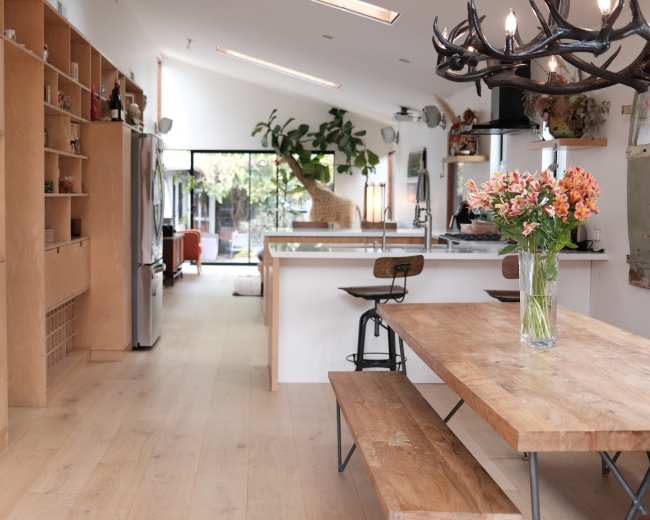 A modern kitchen with wooden cabinetry, a central dining table, and large windows that open to a living area filled with plants and natural light.