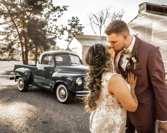 A bride and groom share a kiss in front of a vintage truck parked near a rustic building.