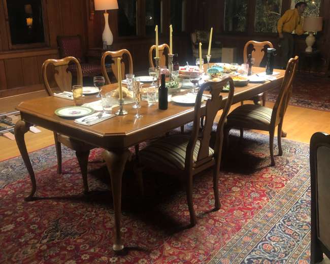 A wooden dining table is set with dishes and candles in a warmly lit room featuring large windows and wooden paneling.