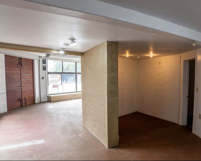 The image shows an empty room with bare walls, a concrete floor, and large windows along one side.