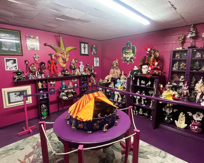 The image shows a vibrant room with pink walls filled with various colorful dolls, masks, and decorative items displayed on shelves and a circular table.