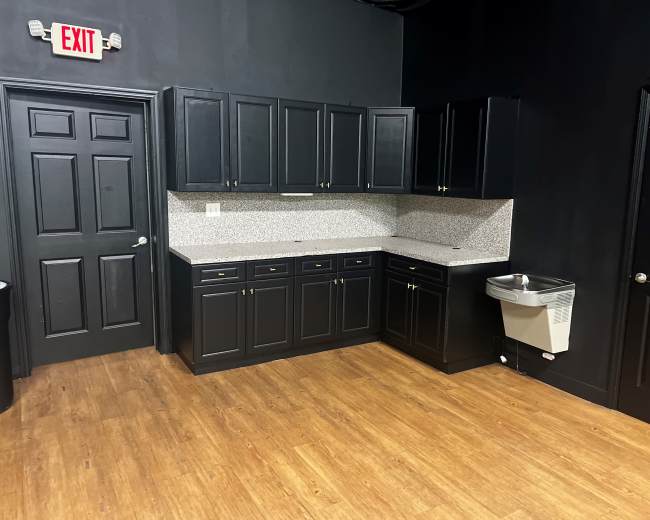 The image shows a kitchen with black cabinetry, a speckled countertop, and a metal sink against a dark wall.