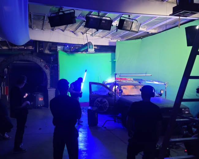 A film crew is working in a studio with a green screen, preparing equipment around a modified vehicle under artificial lighting.