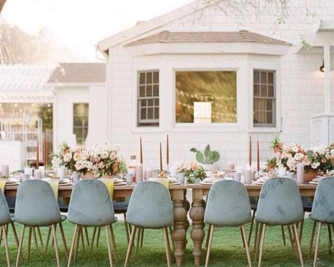 A long table is set with blue chairs and floral arrangements in a backyard near a white house.