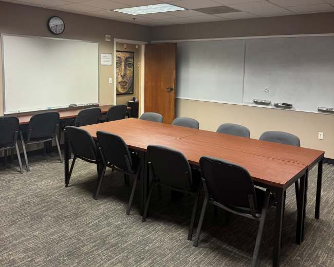 The image shows a conference room with a long wooden table surrounded by black chairs, whiteboards on two walls, and a clock on the back wall.