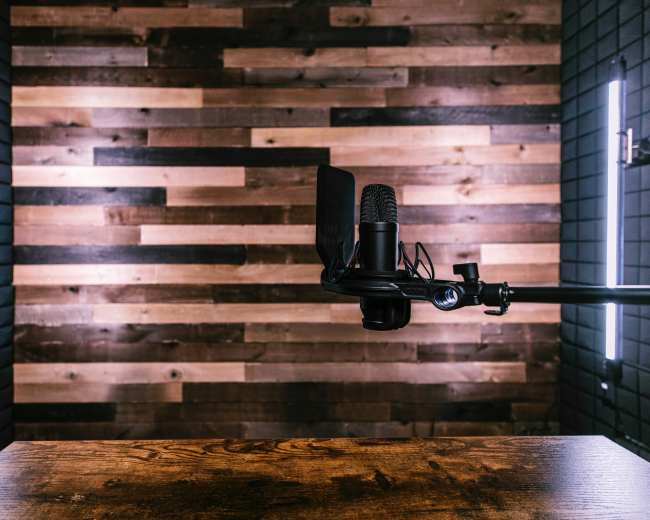 A microphone is positioned on a black arm in front of a wooden table, with a backdrop of wooden planks illuminated by two vertical lights.