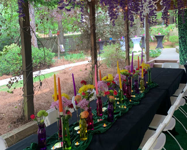 A long table adorned with colorful candles in glass vases is set under a pergola decorated with purple flowers and white chandeliers.