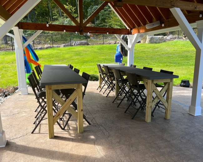 A covered outdoor pavilion features two long wooden tables with chairs arranged around them on a concrete floor.