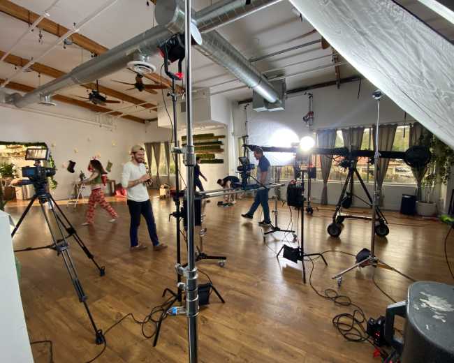A spacious studio is set up for a photoshoot, featuring several cameras and lights, with people preparing in the background.