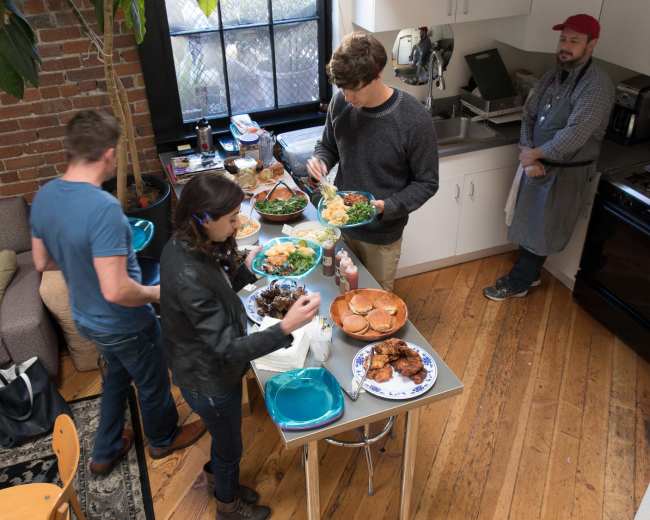 A group of four people are serving themselves food from a buffet-style setup in a kitchen area with a rustic brick wall.
