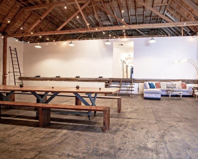 The image shows a spacious interior of a venue featuring wooden beams, a long communal table, a comfortable sofa area, and a staircase leading to another section of the space.