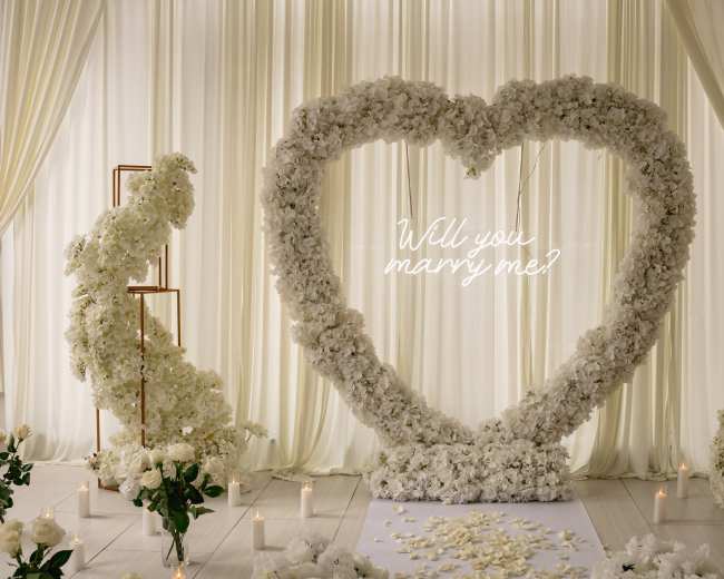 The image features a large heart-shaped floral arrangement with the text "Will you marry me?" displayed in front of a white curtain backdrop.
