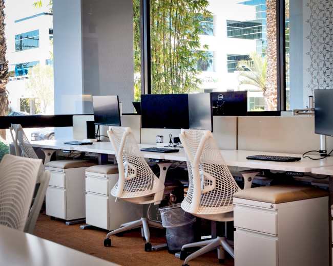 A modern office space with several white ergonomic chairs and computer desks near large windows.