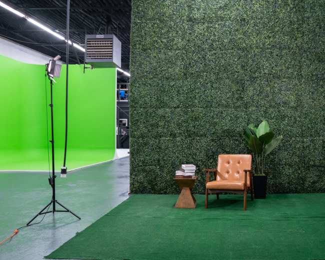 The image shows a photo studio setup featuring a green screen area, an artificial grass floor, a tan armchair, a small wooden side table with books, and a potted plant.
