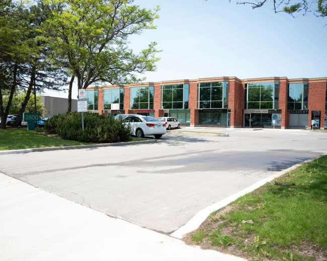 The image shows a parking lot with several cars and a modern brick building featuring large glass windows and greenery around the area.