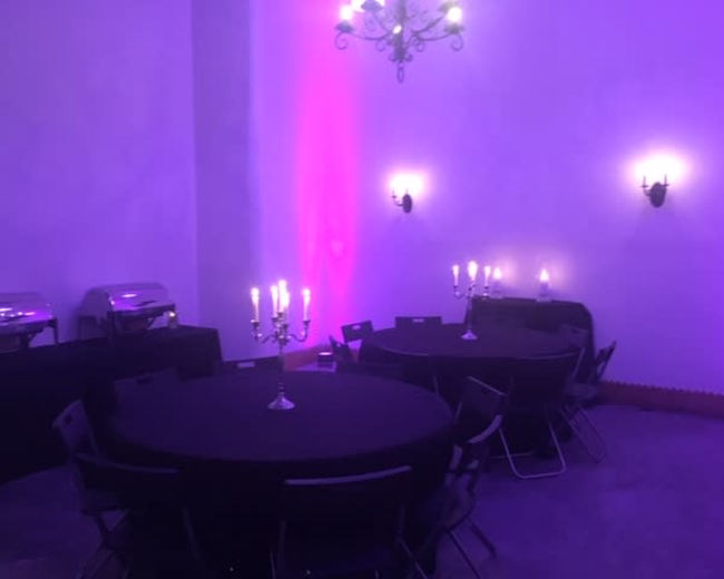 A room with purple lighting features several round tables set with black tablecloths and candelabras, alongside buffet setups against the walls.