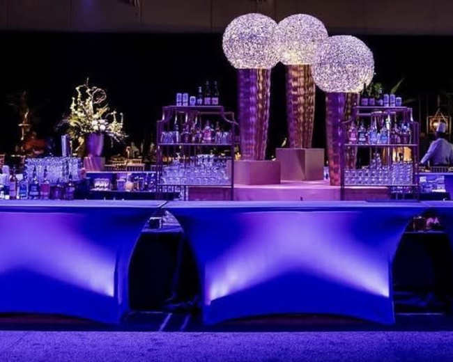 The image shows a elegantly designed bar setup with illuminated tables, floral arrangements, and ornate light fixtures.