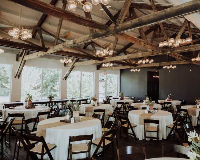 The image shows a spacious event venue with round tables set for dining, wooden beams overhead, and large windows allowing natural light to enter.